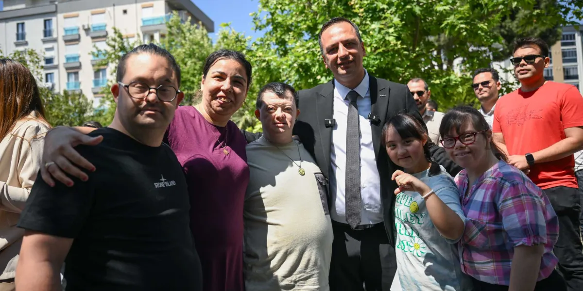 Açık havada, ağaçlıklı bir alanda bir araya gelen bir grup insan, gülümseyerek objektife poz veriyor. Grubun merkezinde, siyah takım elbiseli ve kravatlı bir erkek yer alıyor; etrafında ise Down sendromlu bireyler ve onlara eşlik eden yetişkinler bulunuyor. Katılımcılar samimi bir şekilde yan yana durarak birbirlerine sarılıyor. Arka planda ise yerleşim yerlerine ait binalar görünüyor.
