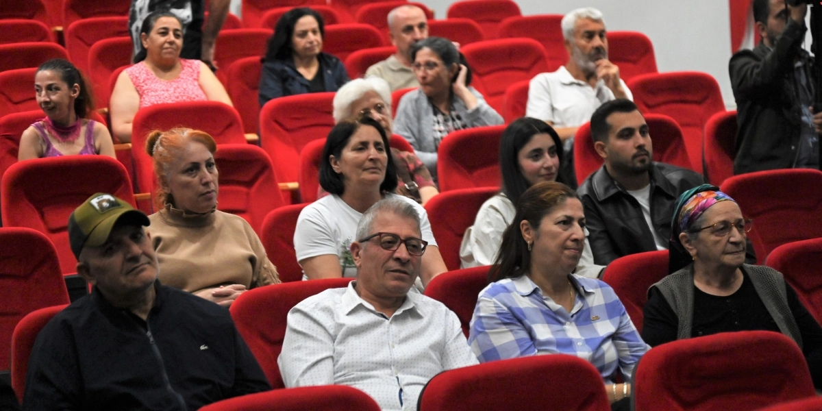 Konferans salonunda kırmızı koltuklarda oturan ve dikkatle sahneye bakan çeşitli yaş gruplarından oluşan bir izleyici grubu. Ön planda gözlüklü bir beyefendi, yanında kareli gömlekli bir kadın ve başında renkli bir eşarp olan yaşlı bir kadın yer alıyor. Arka sıralarda ise farklı kıyafetler içindeki diğer izleyiciler sahneyi izliyor.