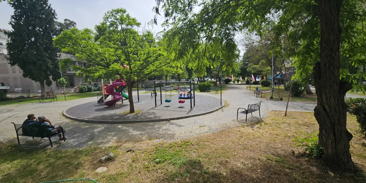 Güneşli bir günde, ağaçlarla çevrili bir parkın içinde yer alan çocuk oyun alanı. Parkın merkezinde, dairesel bir zemin üzerine kurulu mor bir kaydırak, salıncaklar ve dönence bulunuyor. Parkın kenarındaki banklarda oturan iki kişi ve çevredeki yeşil alanlar görülüyor.