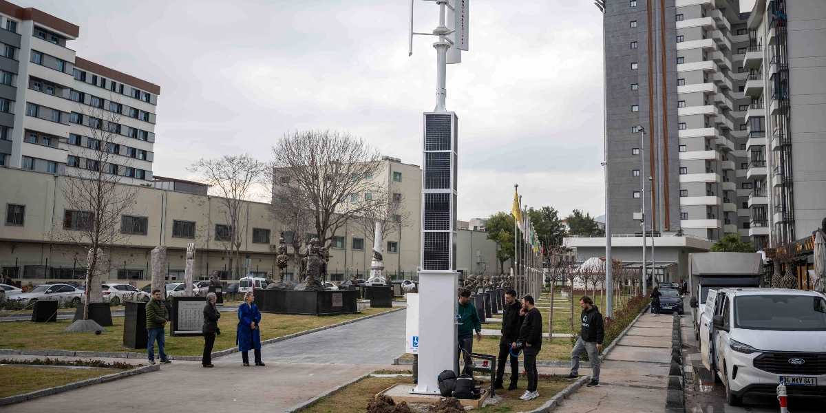 Bornova'da bir park alanında, üzerinde güneş panelleri bulunan dikey eksenli bir rüzgar türbini direği yer alıyor. Direğin çevresinde inceleme yapan bir grup insan bulunurken, arka planda heykellerle süslü bir yürüyüş yolu, ağaçlar, yüksek apartmanlar ve park halindeki araçlar görülüyor.