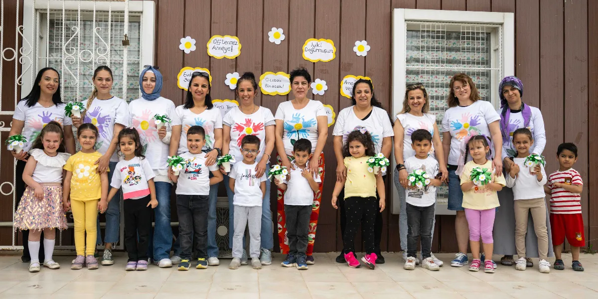 Ahşap kaplamalı bir duvarın önünde yan yana dizilmiş, üzerinde el baskısı desenli beyaz tişörtler giyen bir grup kadın ve çocuk. Arka planda duvara asılmış papatya figürleri ve üzerinde 