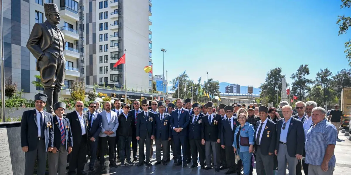 Gaziler Günü anma töreninde, Atatürk heykelinin önünde bir araya gelen gaziler, belediye yetkilileri ve vatandaşlar toplu halde poz veriyor. Aralarında madalyalı gazilerin de bulunduğu grup, açık havada, modern binaların önünde yer alıyor.