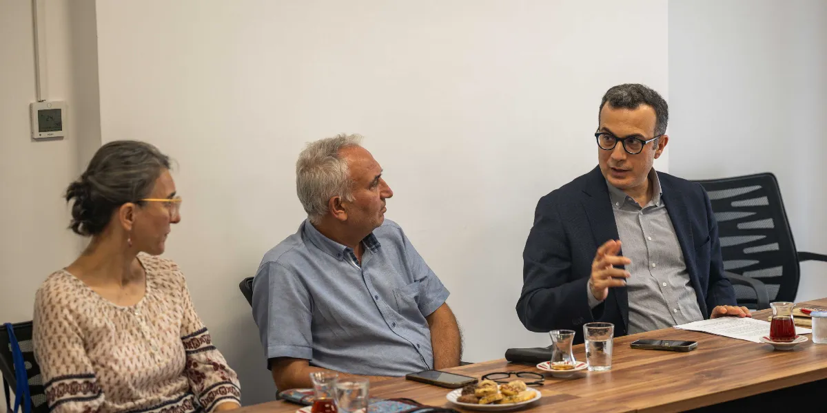 Beyaz bir duvarın önünde, ahşap bir toplantı masasında oturan üç kişi bir konu hakkında konuşuyor. Sağ tarafta lacivert ceketli ve gözlüklü bir erkek, el hareketleriyle bir şeyler anlatırken, yanındaki gri gömlekli orta yaşlı bir erkek ve sol taraftaki desenli bluz giyen kadın onu dikkatle dinliyor. Masanın üzerinde çay bardakları, bir tabak kurabiye ve akıllı telefonlar bulunuyor.