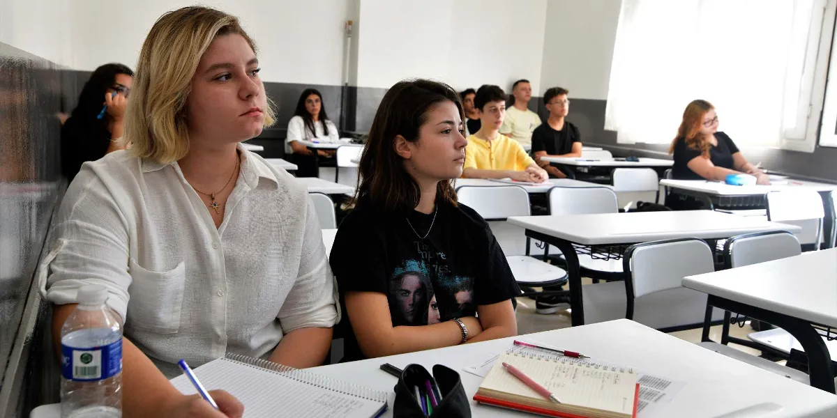 Sınıf ortamında sıralarda oturan öğrenciler dersi dikkatle dinliyor. Ön planda beyaz gömlekli sarışın bir genç kadın ve yanında siyah tişörtlü esmer bir genç kadın masada not alırken görülüyor. Arka planda ise farklı sıralarda oturan diğer öğrenciler yer alıyor. Masaların üzerinde defterler, kalemler ve su şişeleri bulunuyor.