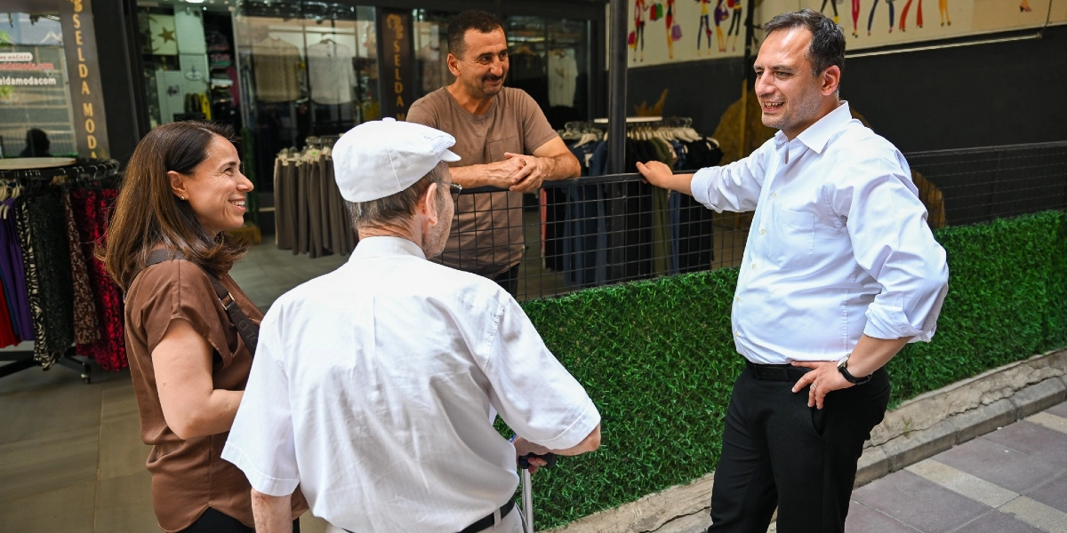 Beyaz gömlekli bir erkek, Çamdibi'ndeki bir giyim mağazasının önünde duran yaşlı bir adam, bir kadın ve bir esnafla samimi bir şekilde sohbet ediyor.
