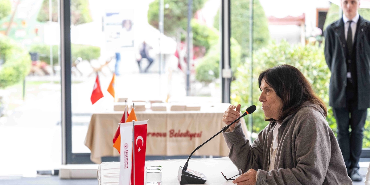 Yazar Latife Tekin, üzerinde Bornova Belediyesi yazılı bir örtünün bulunduğu masada, önündeki mikrofona konuşma yapıyor. Masanın üzerinde küçük bir Türk bayrağı ve Bornova Belediyesi flaması yer alıyor. Arka planda cam bölmeli aydınlık bir salon ve sağ tarafta takım elbiseli bir görevli görülüyor.