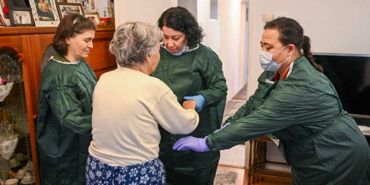 Yeşil koruyucu önlükler giymiş üç sağlık çalışanı, ev ortamında yaşlı bir kadına destek oluyor. Sağlık çalışanlarından ikisi kadının kollarından tutarak ayağa kalkmasına yardımcı olurken, diğeri yanında duruyor. Sağlık çalışanlarının yüzlerinde maske ve ellerinde eldiven bulunuyor.