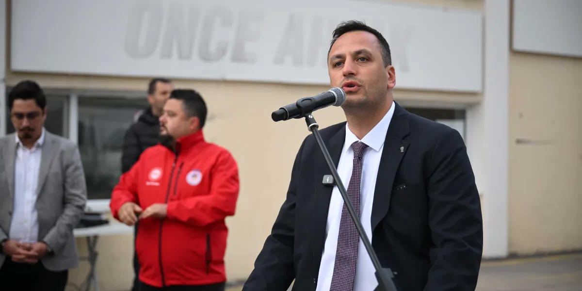 Ömer Eşki, üzerinde beyaz gömlek, desenli kravat ve koyu renkli bir ceketle mikrofon başında konuşma yapıyor. Arka planda, üzerinde kısmen görünen 