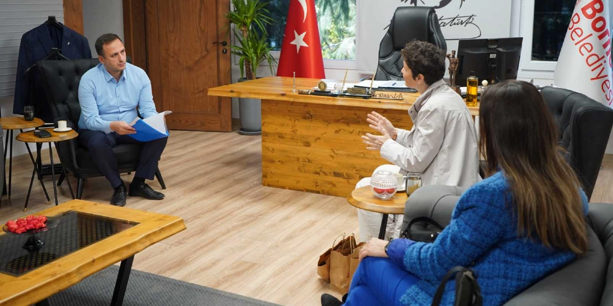 Bornova Belediye Başkanı Ömer Eşki, makam odasında iki kadın konuğuyla bir araya gelerek diyabet dostu menü çalışmaları hakkında görüşme yapıyor. Başkan mavi gömlek ve lacivert pantolonla siyah deri koltukta otururken, karşısındaki konuklar projeyle ilgili detayları aktarıyor. Arka planda Türk bayrağı, Atatürk imzalı bir tablo ve Bornova Belediyesi flaması yer alıyor.