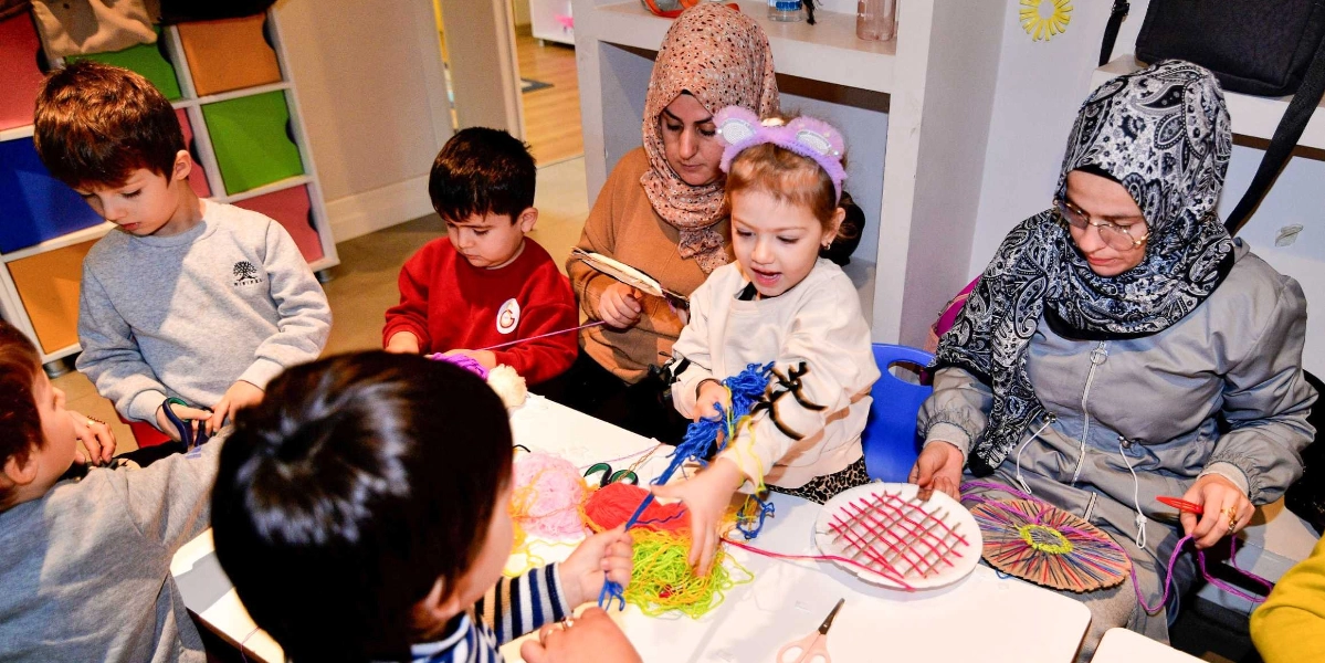 Bir atölye ortamında beyaz bir masa etrafında toplanmış çocuklar ve onlara eşlik eden iki kadın, renkli iplerle el işi çalışmaları yapıyor. Çocuklar masanın üzerindeki yün yumaklarını kullanarak karton tabaklar üzerine örgü desenleri oluşturuyor. Başörtülü kadınlar çocuklara rehberlik ederken, çocuklar dikkatle iplerle uğraşıyor. Arka planda ise renkli çekmeceli bir dolap ve raflar bulunuyor.