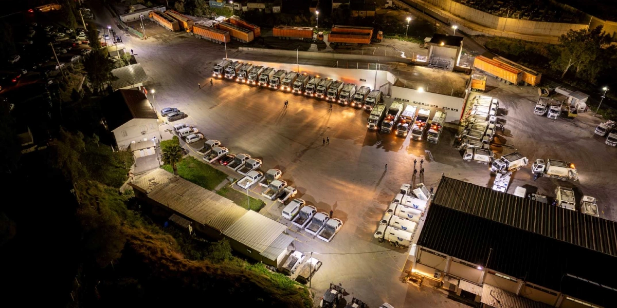 Gece vakti yukarıdan çekilmiş geniş bir şantiye alanında, düzenli sıralar halinde park edilmiş çok sayıda çöp toplama kamyonu ve hizmet aracı görülüyor. Aydınlatılmış geniş bir beton zeminde, bir kısmı yan yana dizilmiş, bir kısmı ise farklı noktalara dağılmış beyaz renkli kamyonlar ve küçük hizmet araçları yer alıyor. Arka planda büyük tır dorseleri ve şantiye binaları bulunurken, alanın genelinde gece mesaisine devam eden veya park halindeki araçların oluşturduğu hareketli bir çalışma ortamı göze çarpıyor.