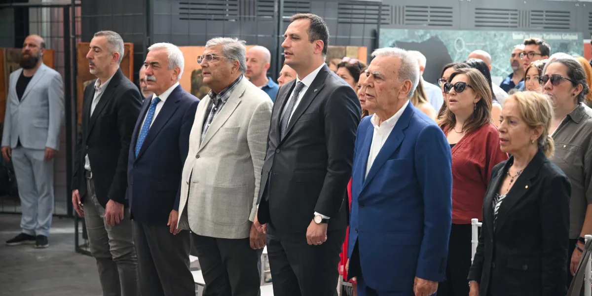 Kapalı bir mekanda, resmi kıyafetli bir grup insan yan yana ayakta durarak saygı duruşunda bulunuyor. Ön sırada takım elbiseli erkekler ve şık giyimli kadınlar yer alıyor; arkada ise daha kalabalık bir grup bulunuyor. Grubun önünde, muhtemelen bir anma törenini temsil eden boş bir koltuk veya alan dikkat çekiyor.
