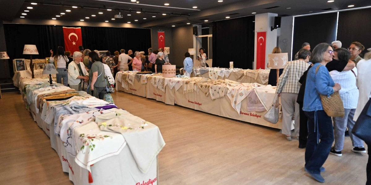 Atatürkçü Düşünce Derneği tarafından düzenlenen, üzerinde el emeği nakışlı örtülerin ve dekoratif ürünlerin sergilendiği uzun masaların yer aldığı bir etkinlik salonu. Bornova Belediyesi logolu örtülerle kaplı masaların etrafında dolaşan ve ürünleri inceleyen katılımcılar görülüyor. Salonun arka duvarında Türk bayrakları asılı ve ortamda çeşitli abajurlar ile sergilenen el işi tekstil ürünleri bulunuyor.