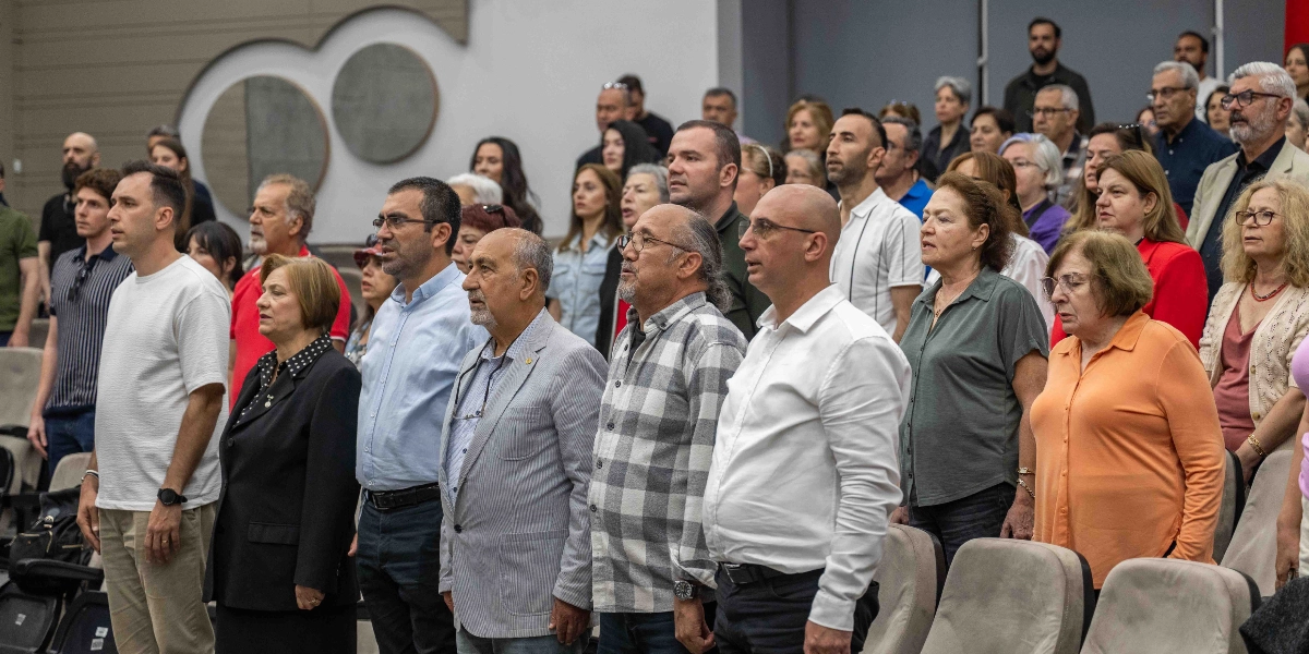 Kapalı bir salonda, ayakta duran ve ciddi bir ifadeyle karşıya bakan, farklı yaş gruplarından oluşan kalabalık bir izleyici grubu. Ön sırada beyaz tişörtlü bir genç, siyah ceketli bir kadın, açık mavi gömlekli bir erkek, gri ceketli ve gözlüklü bir bey, kareli gömlekli bir bey, beyaz gömlekli bir bey, gri bluzlu bir kadın ve turuncu gömlekli bir kadın yan yana dizilmiş şekilde duruyor. Arka sıralarda ise etkinliği takip eden çok sayıda kadın ve erkek izleyici yer alıyor.