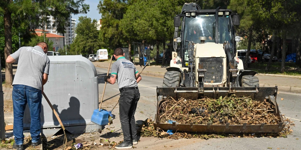 Bornova Belediyesi logolu gri tişörtler giyen iki belediye çalışanı, yol kenarındaki kuru dal ve yaprakları temizliyor. Çalışanlardan biri süpürge ve faraş kullanırken, diğeri bir iş makinesinin kepçesine dolanmış bitki atıklarını düzenliyor. Arka planda ağaçlıklı bir yol ve park halindeki araçlar görülüyor.