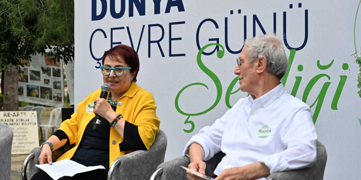 Dünya Çevre Günü etkinliği kapsamında düzenlenen bir panelde, sarı ceketli ve turkuaz gözlüklü bir kadın elinde mikrofonla konuşma yaparken, yanında beyaz gömlekli bir erkek onu dinliyor. Arkalarındaki panoda 