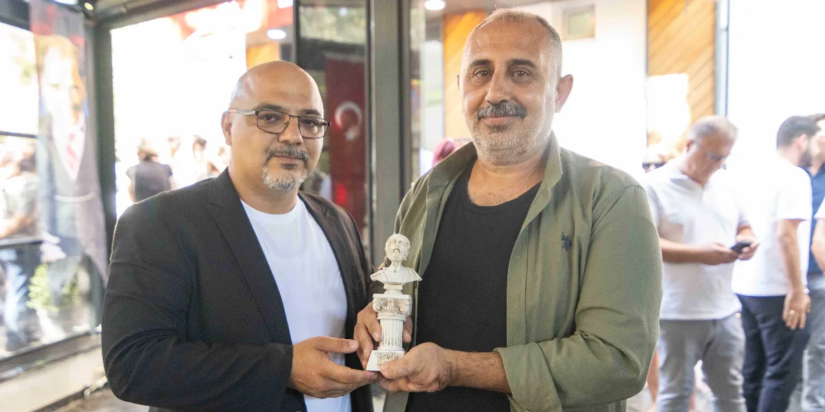 Gözlüklü ve siyah ceketli bir erkek ile haki renkli ceket giyen başka bir erkek, birlikte küçük bir büst heykelini tutuyorlar. Arka planda kalabalık bir ortam ve Türk bayrağı detayı görülüyor.