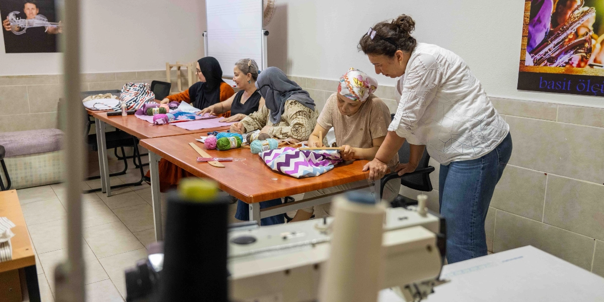 Bornova Belediyesi'nin el sanatları kursunda bir eğitmen, masanın etrafında oturup el işi yapan dört kadına rehberlik ediyor. Kadınların önünde renkli ipler, kumaşlar ve nakış kasnakları bulunuyor. Ön planda ise net olmayan bir dikiş makinesi yer alıyor.