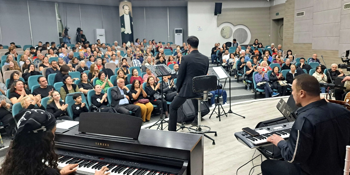 Modern bir konferans salonunda gerçekleşen müzik dinletisinde, sahnede siyah kıyafetli bir erkek solist taburede oturarak şarkı söylüyor. Solistin yanında bir müzisyen klavye çalarken, ön planda piyano başında oturan başka bir müzisyen daha bulunuyor. Salonu dolduran kalabalık izleyici grubu, sahnedeki performansı ilgiyle izliyor ve bazıları alkışlıyor. Salonun arka duvarında büyük bir Mustafa Kemal Atatürk portresi asılı duruyor.