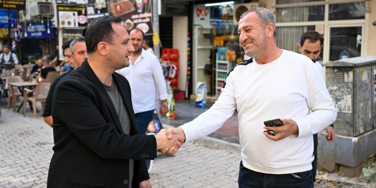 Siyah ceketli bir adam ile beyaz uzun kollu tişört giyen bir adam, Süvari Caddesi'nde yürürken birbirleriyle tokalaşıyor. Beyaz tişörtlü adamın elinde bir cep telefonu bulunuyor ve gülümsüyor. Arka planda dükkanların olduğu hareketli bir cadde ortamı ve çevrede başka insanlar görülüyor.