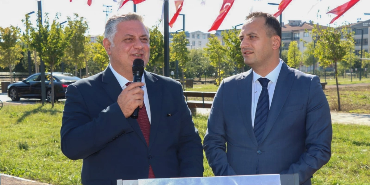 Açık havada, yeşil bir alanda takım elbiseli iki erkek yan yana duruyor. Soldaki kişi elinde mikrofon tutarak konuşma yaparken, sağındaki kişi onu dinliyor. Arka planda ağaçlar, park alanı ve yukarıda asılı Türk bayrakları görülüyor.