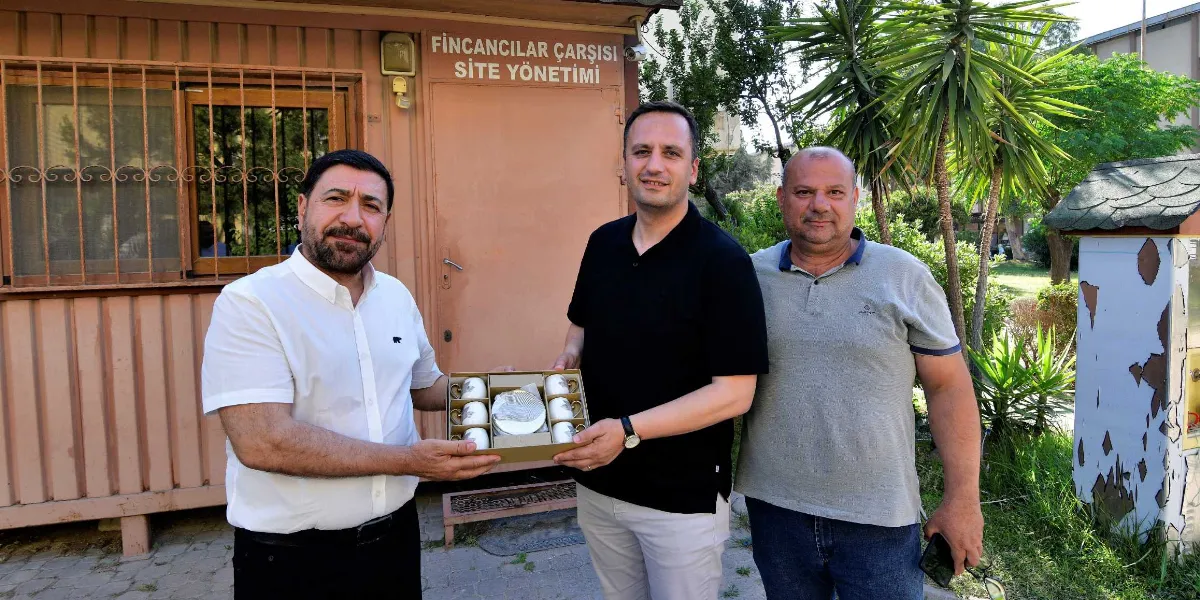 Fincancılar Çarşısı Site Yönetimi binasının önünde duran üç erkek, ortadaki kişinin tuttuğu bir kutu fincan takımını inceliyor. Soldaki kişi beyaz gömlekli, ortadaki kişi siyah polo yaka tişörtlü ve sağdaki kişi gri polo yaka tişörtlüdür. Arka planda yeşil ağaçlar ve bir bahçe alanı görülmektedir.