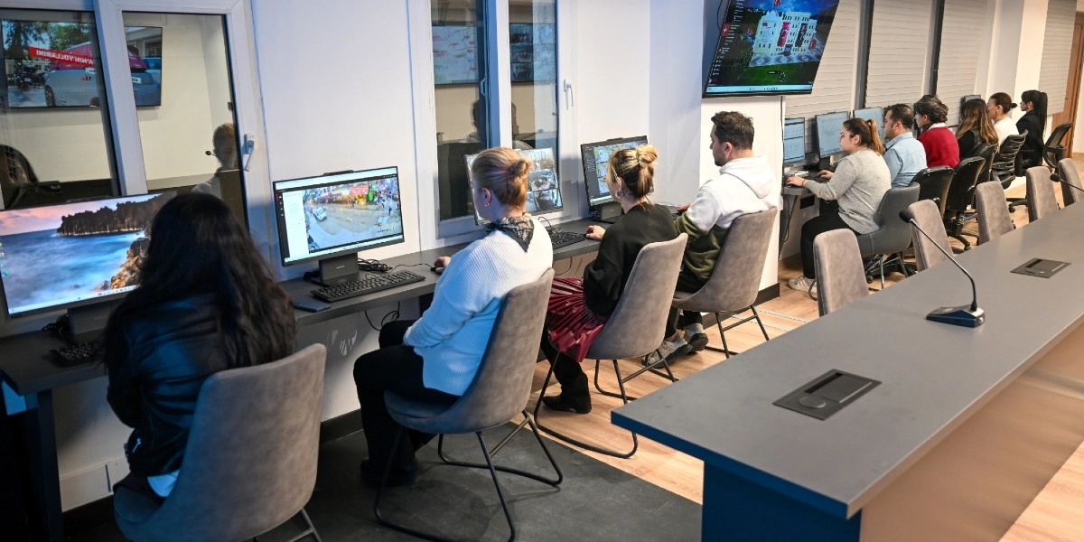 Modern bir ofis ortamında, yan yana dizilmiş bilgisayar masalarında çalışan bir grup insan. Çalışanlar, önlerindeki ekranlarda yer alan görüntüleri ve verileri inceliyor; bazı ekranlarda sokak kameralarından gelen canlı görüntüler, bazılarında ise mimari proje görselleri yer alıyor. Odanın ön kısmında uzun bir toplantı masası ve mikrofon bulunurken, arka planda duvara asılı büyük bir ekranda Bornova'ya ait bir vizyon projenin dijital tasarımı sergileniyor.