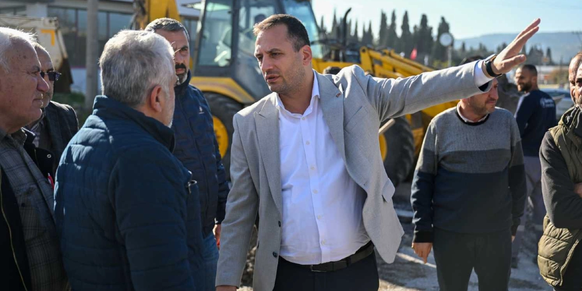 Açık renk ceketli ve beyaz gömlekli bir yetkili, arkasında bir iş makinesinin bulunduğu şantiyede, yanındaki kişilere eliyle bir yönü işaret ederek açıklamalarda bulunuyor. Çevresinde farklı kıyafetler giymiş birkaç erkek daha dikkatle onu dinliyor.