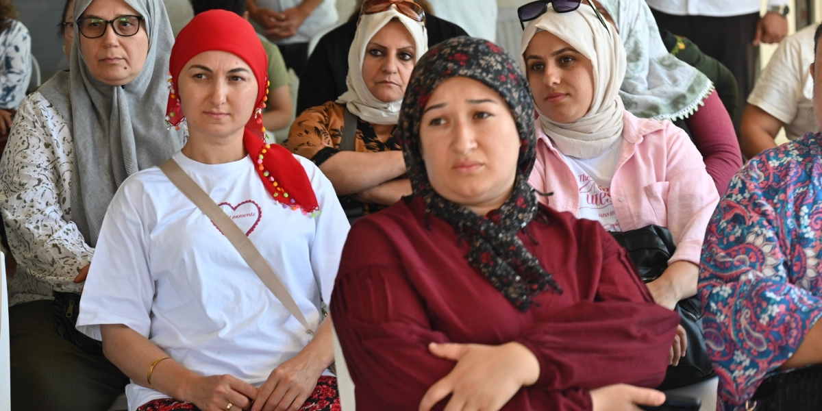 Bir salonda yan yana oturmuş, dikkatle bir noktaya bakan, başörtülü kadınlardan oluşan bir grup. Ön planda, üzerinde beyaz tişört ve kırmızı başörtüsü olan bir kadın ile bordo renkli bluz ve desenli başörtüsü takan başka bir kadın yer alıyor. Arka planda ise farklı renklerde başörtüleri ve kıyafetleri olan diğer katılımcılar bulunuyor.