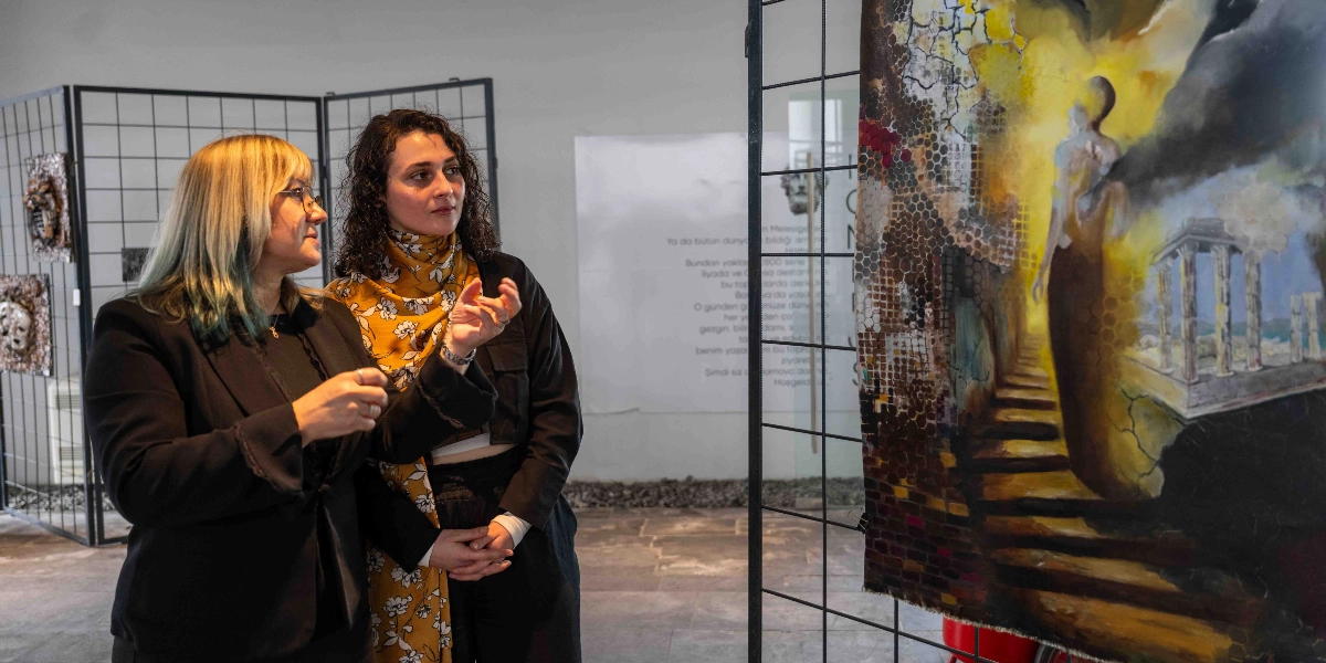 Sanat galerisinde, siyah ceketli sarışın bir kadın ile siyah ceket ve sarı çiçek desenli şal giyen kıvırcık saçlı bir kadın, metal bir ızgara üzerine asılmış büyük boyutlu, antik sütunlar ve soyut figürler içeren bir tabloyu inceliyor. Kıvırcık saçlı kadın el hareketleriyle tablo hakkında konuşurken, yanındaki kadın onu dikkatle dinliyor. Arka planda duvarda metin içeren bir pano ve başka sanat eserleri yer alıyor.
