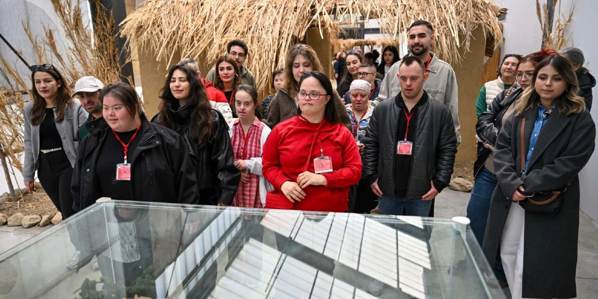 İçlerinde Down sendromlu bireylerin de bulunduğu bir grup genç, müze ortamında cam bir vitrinin etrafında toplanmış, sergilenen eserleri ilgiyle inceliyor. Gençlerin birçoğunun boynunda etkinlik kartı asılı. Arka planda ise sazdan çatılı, kerpiç görünümlü geleneksel bir yapı maketi yer alıyor.