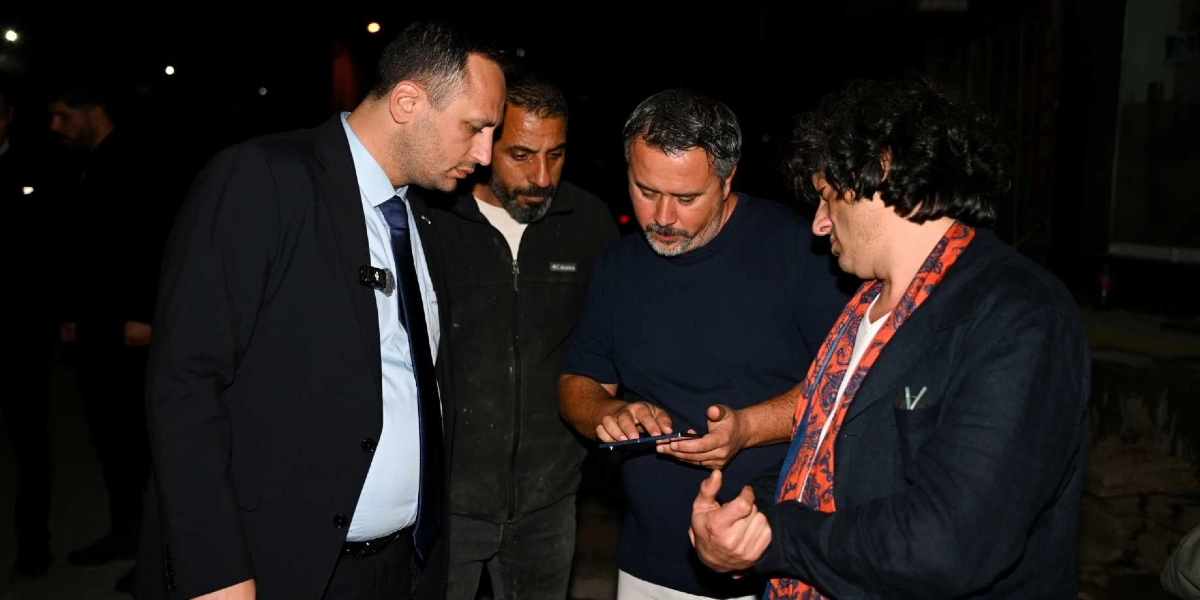 Gece vakti dışarıda bir araya gelen dört erkek, ellerindeki akıllı telefona odaklanmış bir şekilde bir konuyu inceliyor. En solda takım elbiseli ve kravatlı bir kişi, yanında koyu renkli bir ceket giyen sakallı bir adam, ortada lacivert tişörtlü bir kişi ve en sağda ise üzerinde desenli turuncu bir şal bulunan koyu renkli ceketli bir adam yer alıyor.