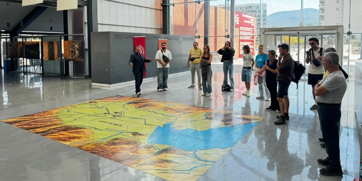 Modern bir müze veya sergi salonunun zeminine yerleştirilmiş, İzmir Körfezi ve çevresini gösteren büyük ölçekli bir haritayı inceleyen bir grup insan. Grubun önünde duran bir rehber, harita üzerinde anlatım yaparken, diğer katılımcılar onu dinliyor. Salonun arka planında Bornova Belediyesi'ne ait bayraklar, duvarlarda sergilenen sanat eserleri ve dışarıyı gösteren geniş cam cepheler bulunuyor.