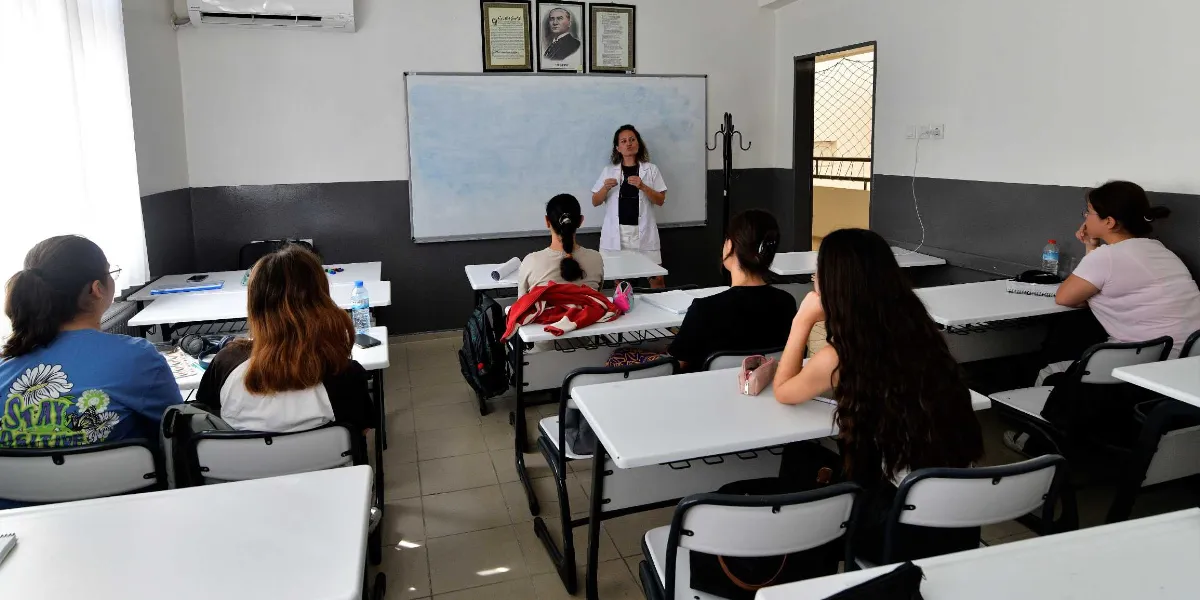 Sınıf ortamında beyaz önlüklü bir eğitmen, tahtanın önünde durarak öğrencilere ders anlatıyor. Öğrenciler sıralarda oturmuş, eğitmeni dinliyor. Tahtanın üzerinde Atatürk portresi ve çerçeveli belgeler asılı.