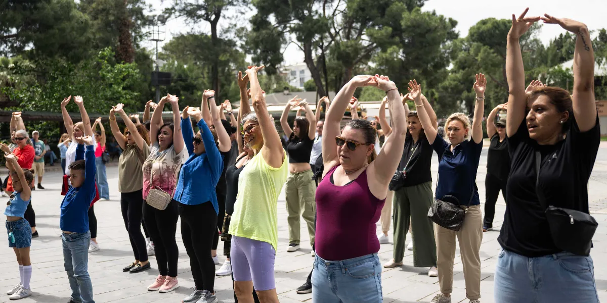 Açık havada, park gibi bir alanda bir araya gelmiş kadınlar ve çocuklar, ellerini havaya kaldırarak hep birlikte esneme hareketleri yapıyor. Farklı yaş gruplarından oluşan grup, spor kıyafetleri içinde ve neşeli bir atmosferde egzersiz yapıyor.
