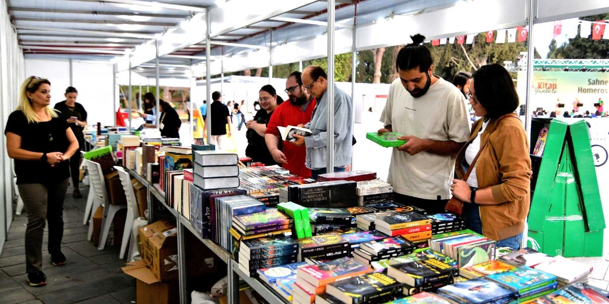 Bornova'da düzenlenen bir kitap fuarı alanında, uzun bir stant masasının etrafında toplanmış insanlar kitapları inceliyor. Ön planda, üzerinde topuz saçlı bir genç adamın elinde yeşil kapaklı bir kitap tuttuğu ve yanında kahverengi ceketli bir kadının kitaplara baktığı görülüyor. Masanın diğer tarafında ise gözlüklü, gri ceketli bir beyefendi ve kırmızı tişörtlü bir kişi kitapları dikkatle inceliyor. Arka planda ise stant görevlisi olduğu anlaşılan sarışın bir kadın ayakta bekliyor. Fuarda kitapların dizili olduğu masaların yanı sıra, arka planda 