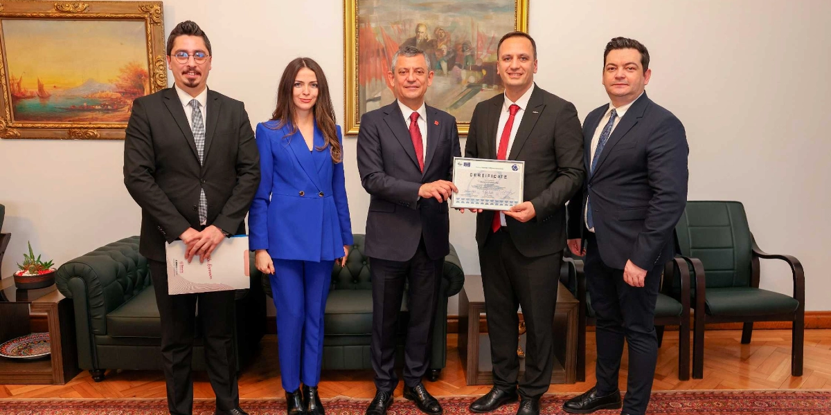 Beş kişilik bir grup, resmi bir toplantı ortamında yan yana durarak poz veriyor. Gruptakilerin çoğu takım elbiseli, ortadaki iki kişi ise Avrupa Konseyi tarafından verilen bir başarı sertifikasını birlikte tutuyor. Arka planda klasik tabloların asılı olduğu krem rengi duvarlar ve koyu yeşil deri koltuklar bulunuyor.