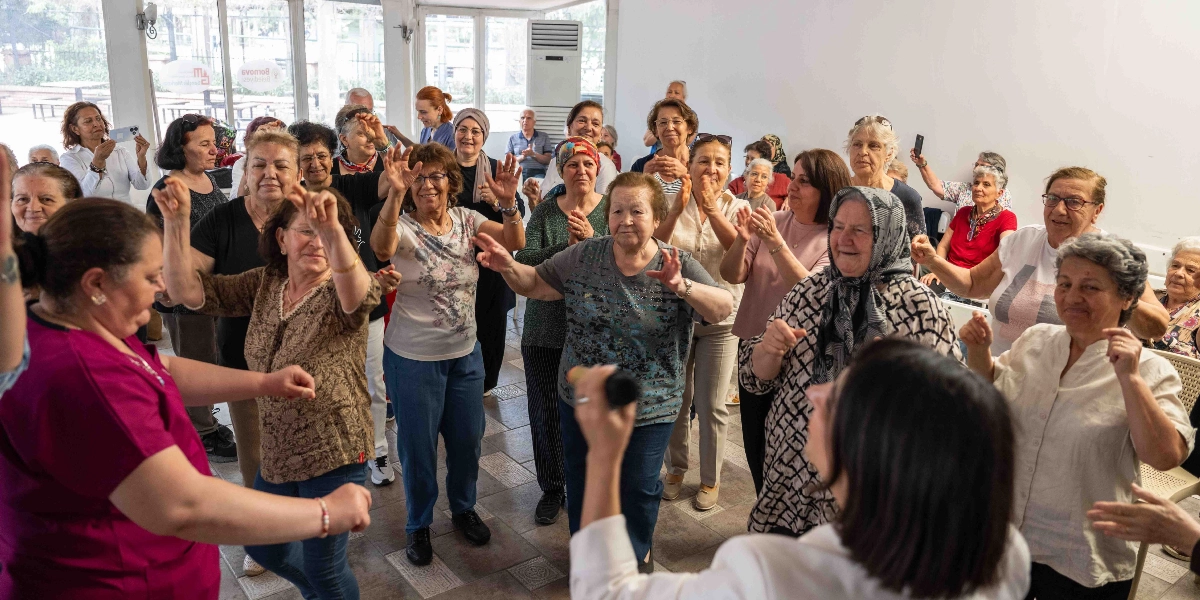 Aydınlık ve geniş bir salonda toplanmış, çoğunluğu yaşlı kadınlardan oluşan bir grup, müzik eşliğinde neşeyle dans edip ellerini çırpıyor. Ön planda sırtı dönük bir kişi elinde mikrofonla şarkı söylerken, diğer katılımcılar günlük kıyafetleriyle bu coşkuya eşlik ediyor. Arka planda ise Bornova Belediyesi logosunun yer aldığı cam bölmeler ve içerideki hareketliliği izleyen diğer katılımcılar görülüyor.