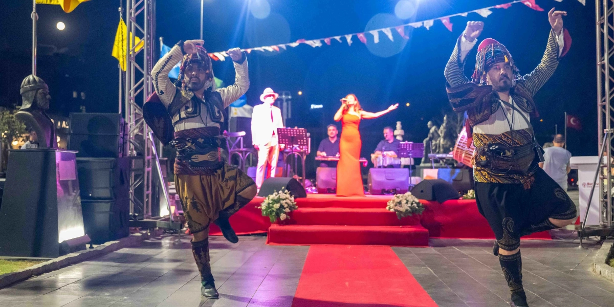 Gece düzenlenen bir açık hava etkinliğinde, geleneksel kıyafetler giymiş iki zeybek dansçısı sahnede tek ayak üzerinde figür sergiliyor. Arka planda kırmızı halı kaplı sahnede, kırmızı elbiseli bir kadın şarkıcı mikrofonla şarkı söylerken, ona enstrümanlarıyla eşlik eden müzisyenler bulunuyor. Sahne alanı Türk bayrakları ve ışıklandırmalarla süslenmiş durumda.