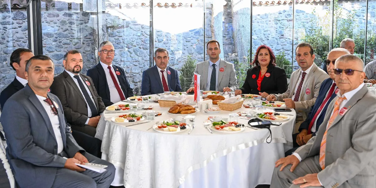 Taş duvarlı ve cam bölmeli bir mekanda, beyaz örtülü yuvarlak bir masa etrafında toplanmış, yakalarında Bornova Muhtarlar Akademisi logolu rozetler bulunan bir grup kadın ve erkek muhtar kahvaltı yapıyor. Masada çeşitli kahvaltılıklar, ekmek sepetleri ve küçük bir Türk bayrağı yer alıyor. Katılımcılar resmi kıyafetleriyle kameraya bakıyor.
