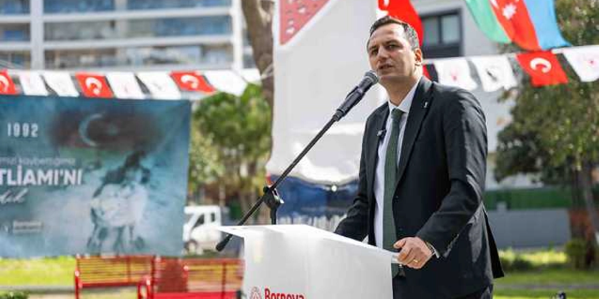 Koyu renk takım elbiseli ve yeşil kravatlı bir erkek, Bornova Belediyesi logolu bir kürsüde mikrofon başında konuşma yapıyor. Arka planda, üzerinde 