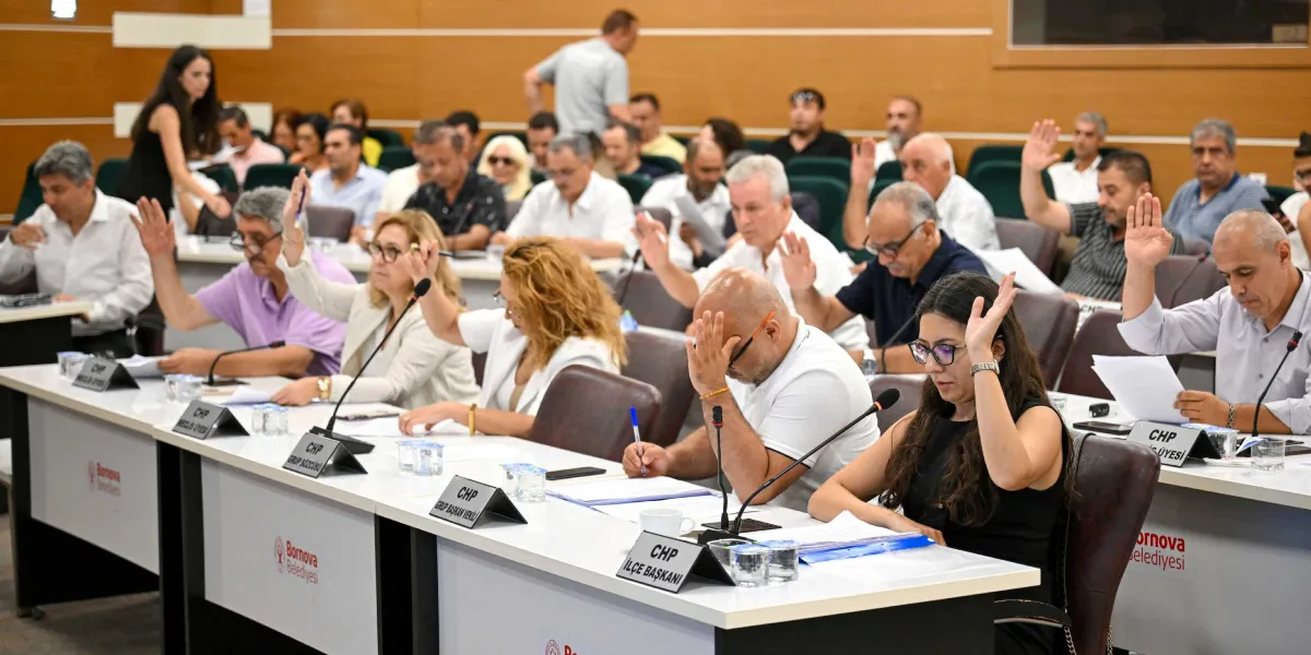 Bornova Belediyesi meclis salonunda, masalarda oturan çok sayıda katılımcının bir oylama sırasında ellerini kaldırarak oy kullandığı toplantı anı. Ön planda, üzerinde 