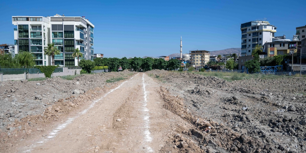 Şehir merkezinde, üzerinde beyaz kireçle çizilmiş hatların bulunduğu toprak bir yol ve çevresinde yeni yapılmış apartmanlar, ağaçlık alanlar ve uzaktan görünen bir cami minaresi yer alıyor.