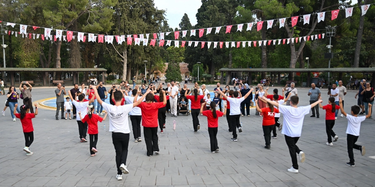 Ağaçlıklı geniş bir meydanda, kırmızı beyaz tişörtlü bir grup çocuk ve genç, ellerini havaya kaldırarak halk oyunu sergiliyor. Meydanın üzerinde Türk bayrakları ve Atatürk görselleriyle süslü flamalar asılı. Çevredeki yetişkinler ise bu gösteriyi izliyor ve bazıları cep telefonlarıyla kayıt alıyor.