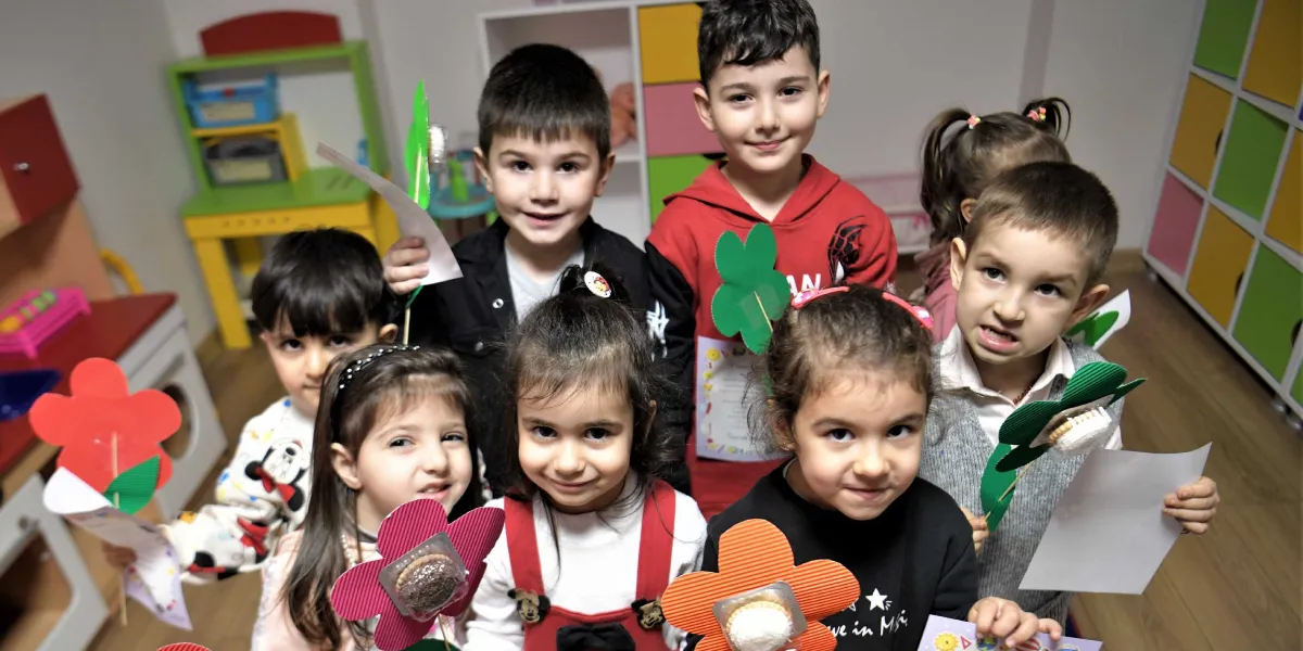 Çocuk oyun merkezinde ellerinde kartondan yaptıkları çiçek figürleri ve bisküvilerle poz veren, gülümseyen bir grup çocuk. Arka planda renkli dolaplar ve oyuncak mutfak ünitesi bulunan bir sınıf ortamı görülüyor.