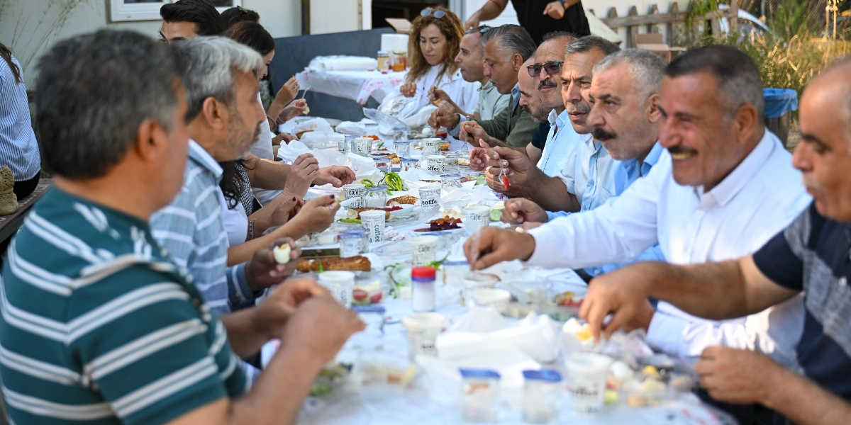 Açık havada, uzun bir masanın etrafında toplanmış kalabalık bir grup insan kahvaltı yapıyor. Masanın üzerinde çeşitli kahvaltılıklar, küçük plastik bardaklar ve tabaklar bulunuyor. Birbirleriyle sohbet eden ve gülümseyen insanlar, dayanışma içinde birlikte yemek yiyor.