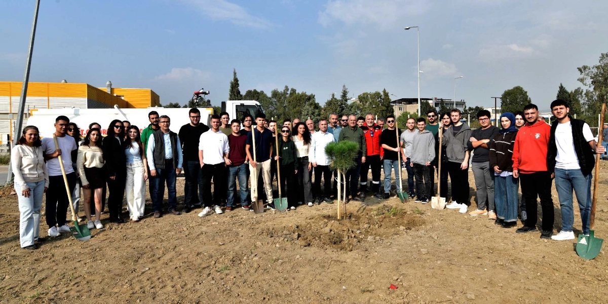 Açık havada, toprak bir alanda bir araya gelmiş kalabalık bir genç grubu ve yetişkinler, yeni dikilmiş küçük bir çam fidanının etrafında duruyor. Katılımcıların birçoğu ellerinde ağaç dikiminde kullanılan kürekler tutuyor. Arka planda beyaz bir araç ve sarı renkli bir bina yapısı görülüyor. Gün ışığının hakim olduğu ortamda grup, hep birlikte hatıra fotoğrafı çektiriyor.