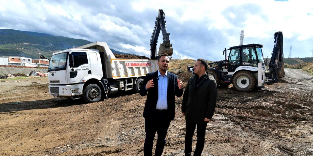 Açık havada, çamurlu bir arazide duran iki erkek, Bornova Belediyesi'ne ait bir damperli kamyon ve bir iş makinesinin önünde konuşuyor. Arka planda, üzerinde 