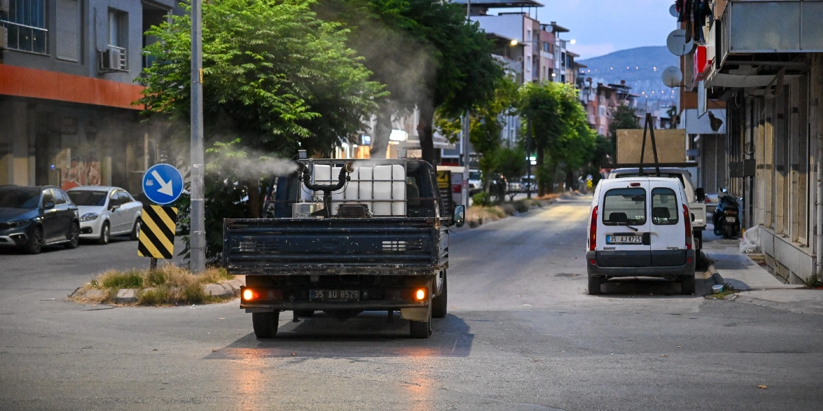 Çamdibi sokaklarında ilaçlama yapan, arkasında beyaz bir tank bulunan 35 AU 8529 plakalı kamyonet, etrafa duman yayarak ilerliyor. Yolun sağ tarafında park halindeki beyaz bir hafif ticari araç ve çevrede çok katlı apartmanlar ile ağaçlar görülüyor.