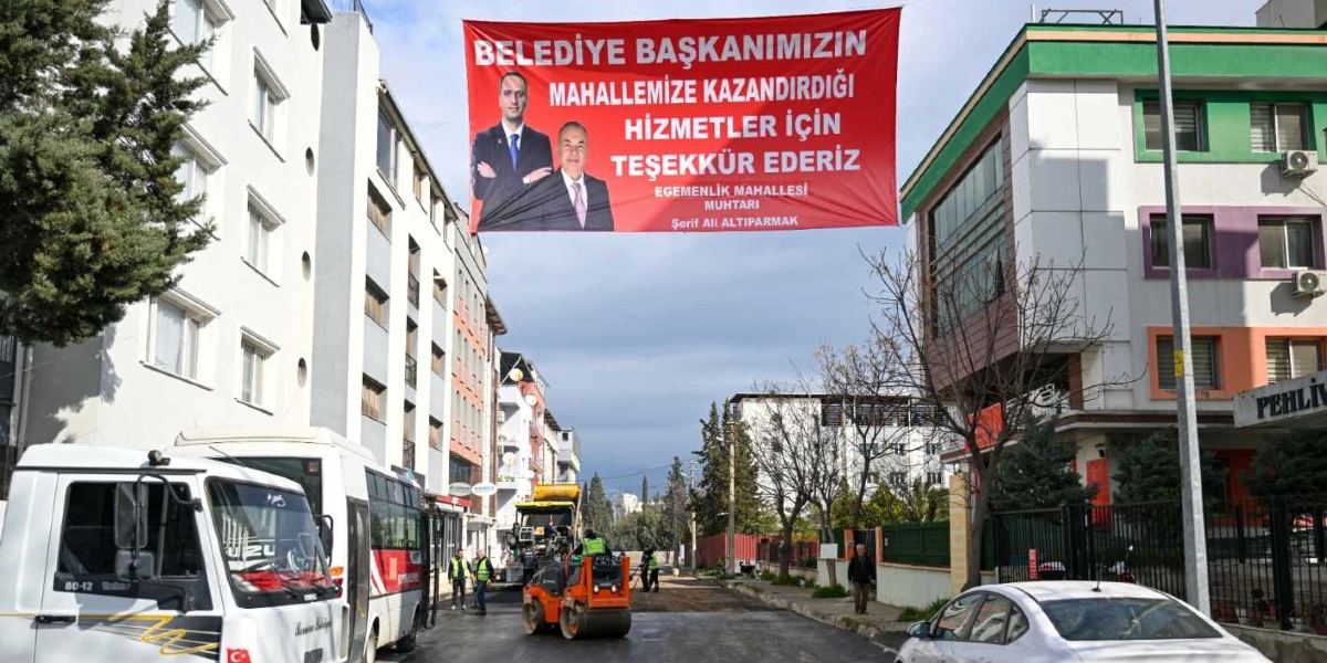 Egemenlik Mahallesi'ndeki bir sokakta, yol yenileme çalışması yapan iş makineleri ve çalışanlar görülüyor. Sokağın üzerinde asılı duran büyük kırmızı pankartta, iki kişinin fotoğrafı ile birlikte 